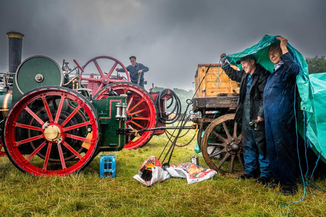 John Appleby, of Pickering, works on  Old Lass, a 1901 7 horsepower agricultural Traction Engine, whilst Mark Angusm and Rod Lytton, shelter from a heavy rain shower.