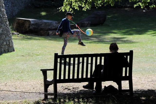 A boy enjoys a ball game in the sun while another person makes the most of the warm weather but stays shaded in Museum Gardens, York.