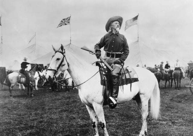 William 'Buffalo Bill' Cody (1846 - 1917) American entertainer, sitting on horseback and holding a rifle, looks off into the distance as British and American flags fly around him. Tents for his Wild West show are in the background.  (Photo by Hulton Archive/Getty Images)