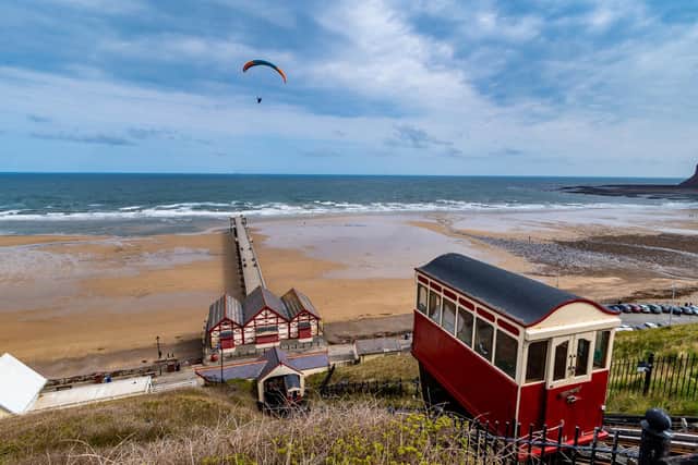 A paraglider flies high above the famous Saltburn Cliff Tramway, a funicular railway system of two counterbalanced cars attached at the end of a long cable that goes from one car, up the slope, around a pulley, and back down to another car. This means that as one car goes up the slope, the other one must go down