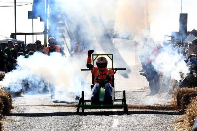 A soapbox racer triumphantly raises their fist in the air as they whizz down the race track with smoke engulfing the air around them.