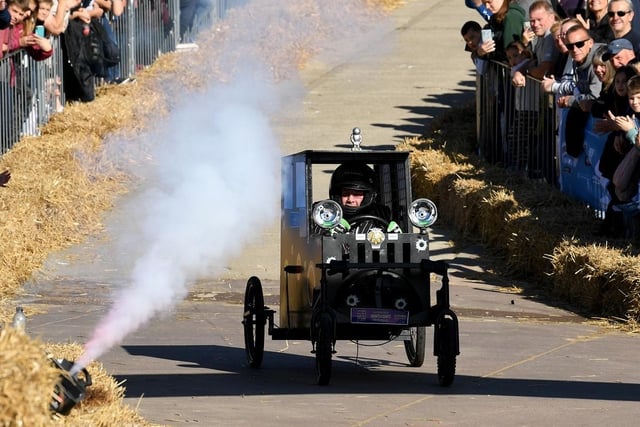 A black soapbox car drives along the race track with smoke blowing in their direction.