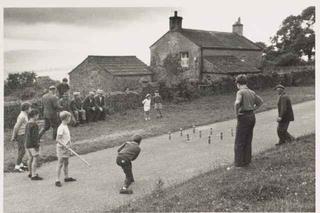 Game of wallops Photographed by Kissling, Werner, June-August 1964 Children playing wallops, or nine-pins, in the street at Castle Bolton (Wensleydale). The game is played by throwing a stick from a distance at nine wooden skittles/pins set out in square. The object of the game is to knock down all nine skittles in as few throws as possible. A group of men can be seen sitting on a bench, watching men from the village playing quoits on the grass verge. Leeds University Library, Special Collections, [LAVC/PHO/P1748]